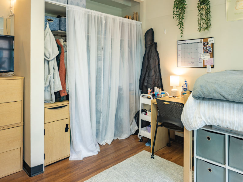 A student dorm room with a bed, desk, open closet, and storage units.
