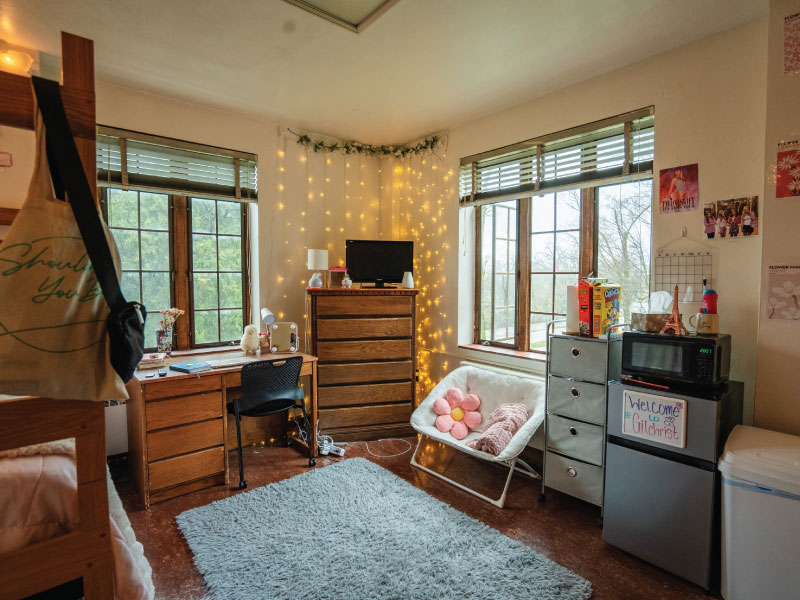 Cozy dorm room with a large window, comfy chair, microwave, fridge, desk, and decorated walls.