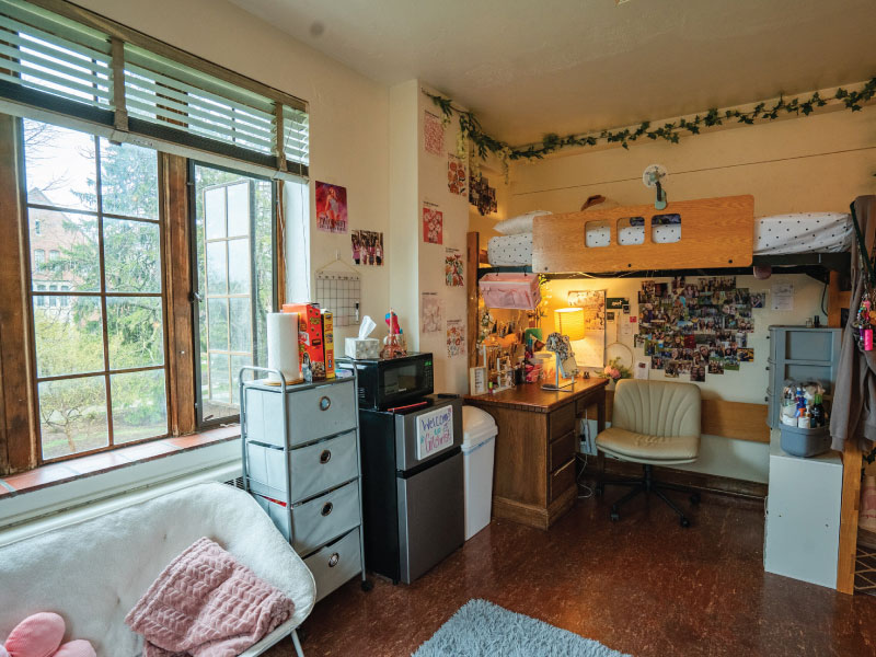 Cozy dorm room with a large window, comfy chair, microwave, fridge, desk, and decorated walls.