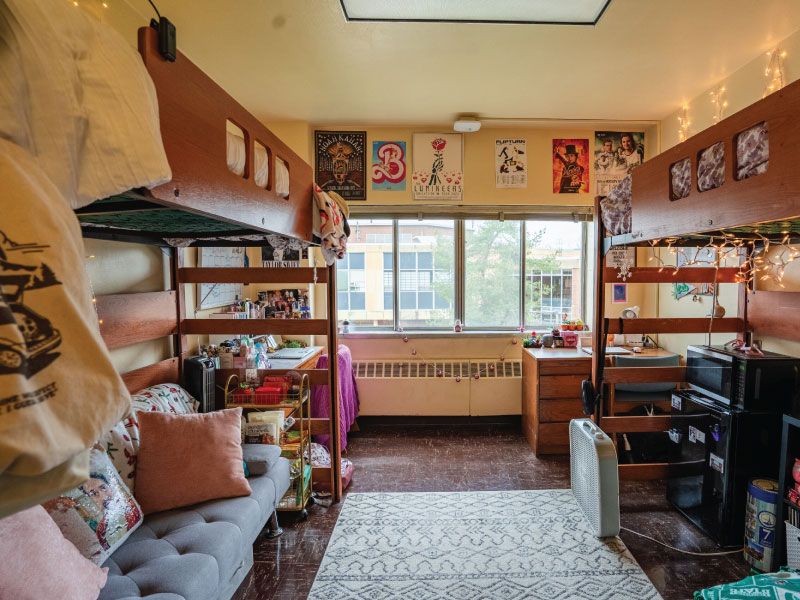 Interior of a dorm room with loft beds, a sofa, desk, mini-fridge, and decorative lights.