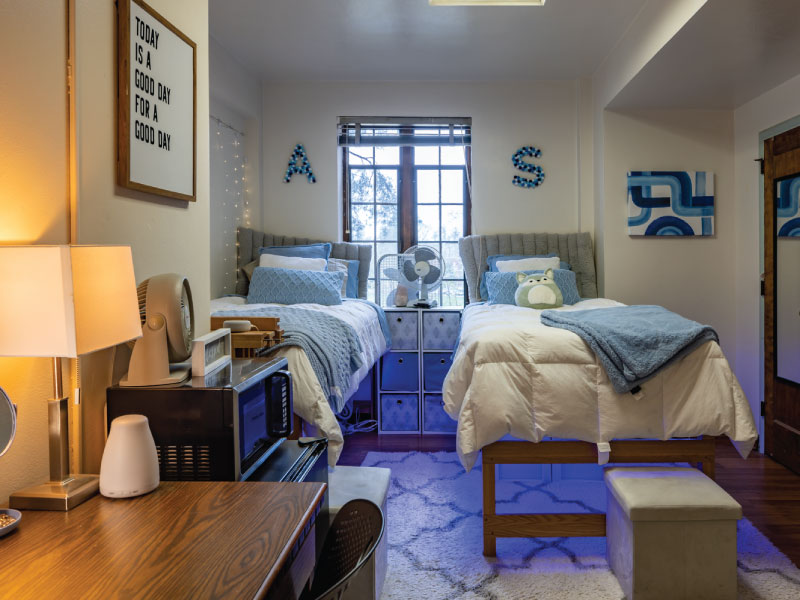 Neatly organized dorm room with twin beds, light blue decor, a window, and a desk with a lamp.