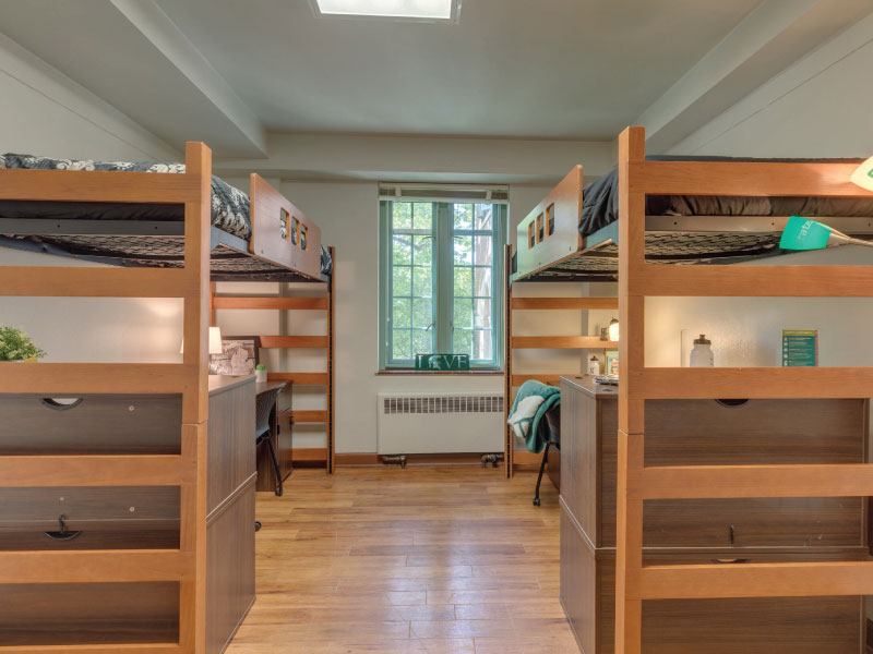 A tidy dorm room with two lofted beds, desks beneath, and a central window allowing natural light.