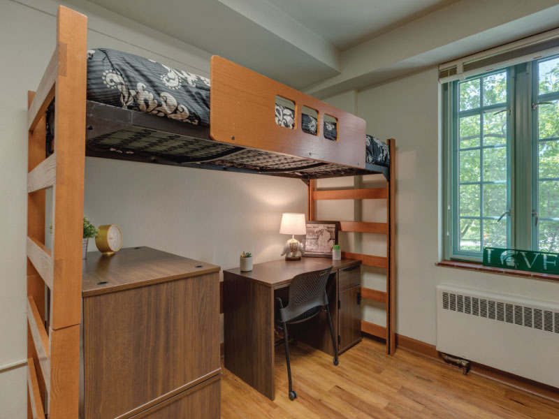A tidy dorm room with two lofted beds, desks beneath, and a central window allowing natural light.